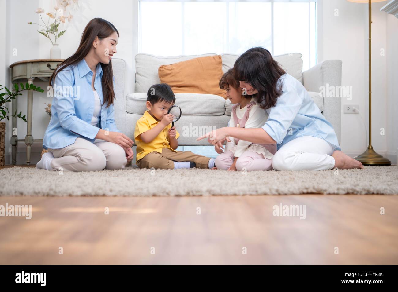 Mutter und Kinder, die sich in einer warmen häuslichen Aktivität beschäftigen, wobei ein Kind eine Lupe benutzt, um Objekte zu erkunden, was das frühe Lernen und Denken fördert Stockfoto