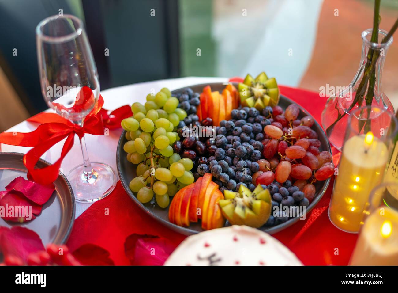 Frischer Obstteller auf romantischem rotem Tisch. Frischer Obstteller mit Trauben, Blaubeeren, Kiwi und geschnittenem Obst auf einem romantischen roten Tisch mit Kerzen Stockfoto