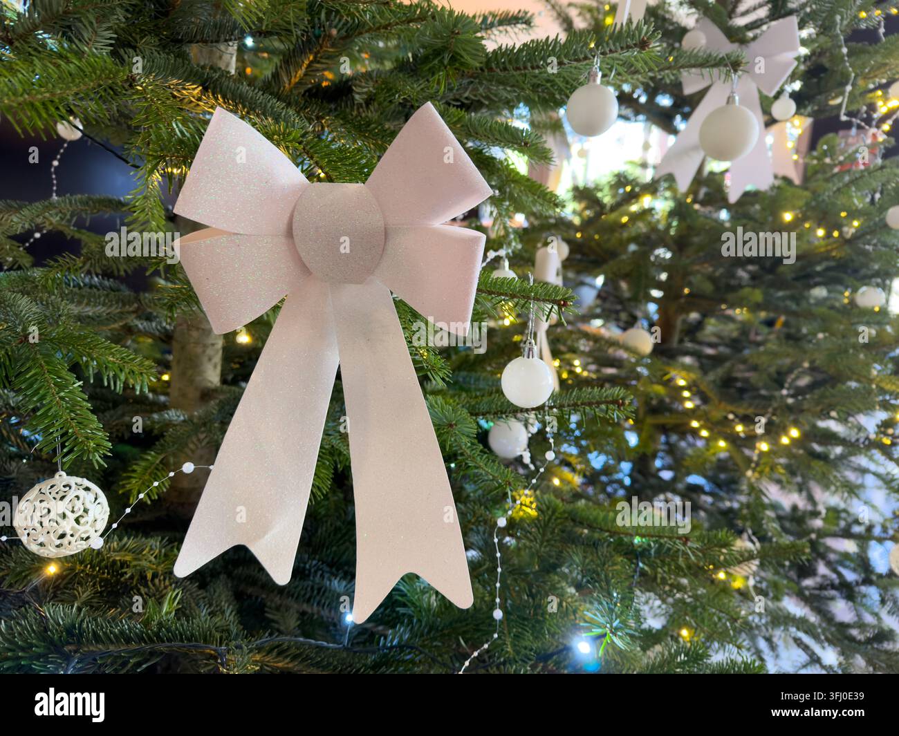 Eine elegante weiße Schleife sitzt auf einem lebendigen Fichtenzweig, der mit leuchtenden Glühbirnen und hängenden Kugeln gefüllt ist, um eine friedliche Winteratmosphäre zu schaffen während der Stockfoto
