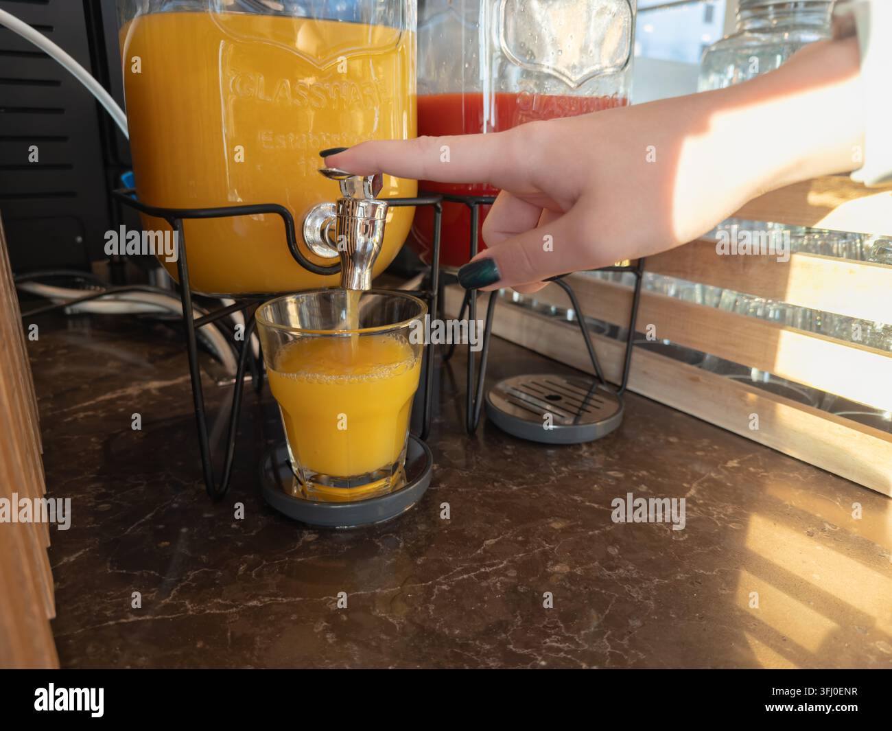 Nahaufnahme einer Hand, die einen Metallhahn drückt, um kalten Orangensaft in ein Glastumbler zu geben, und damit eine morgendliche Frühstücksroutine in einem hellen, modernen Stockfoto