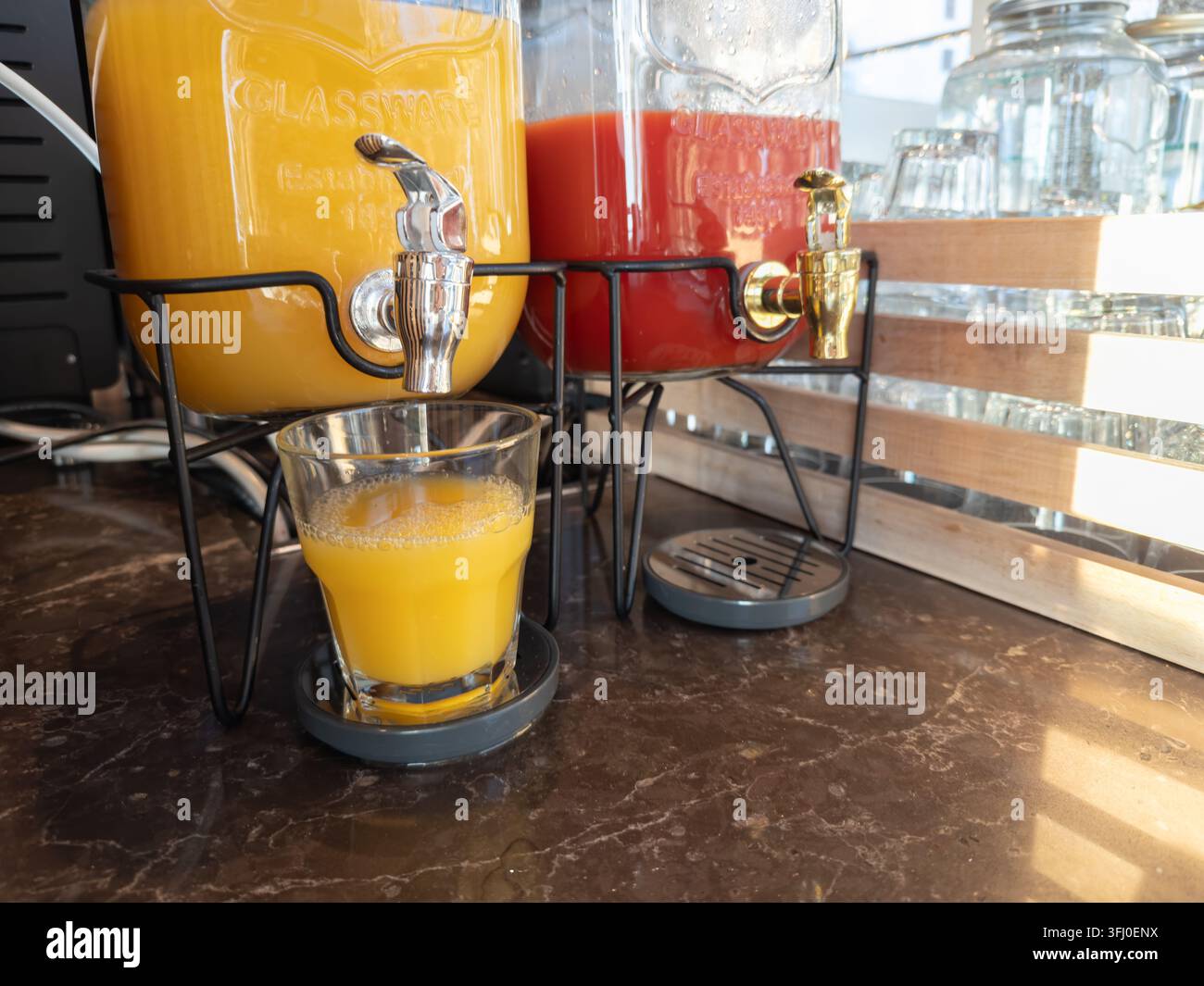 Ein Glas Orangensaft, das aus einem im Vintage-Stil gehaltenen Spender auf einer Marmorarbeitsplatte gegossen wird, reflektiert einen sauberen Morgenfrühstücksservice für Gäste, die suchen Stockfoto