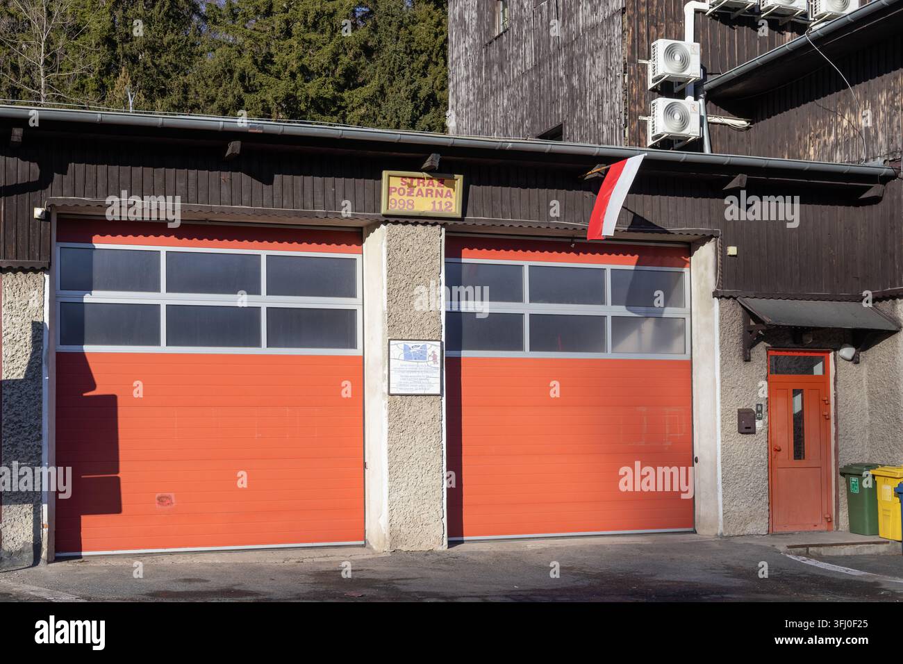 Szklarska Poręba, Polen - 18. Dezember 2025: Die rustikale Fassade eines örtlichen Feuerwehrgebäudes zeigt große orangefarbene Einfahrtstore und eine nationale Fl Stockfoto