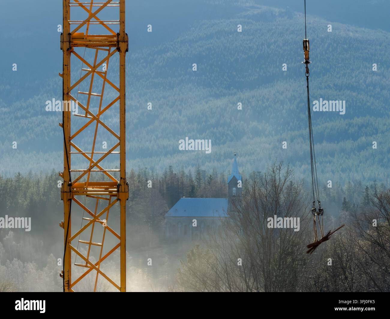 Ein Industriekran steht vor einer friedlichen Kirche, die in einem nebligen Waldgebiet liegt und die städtische Entwicklung symbolisiert, die die traditionelle ruhige Landschaft verwandelt Stockfoto