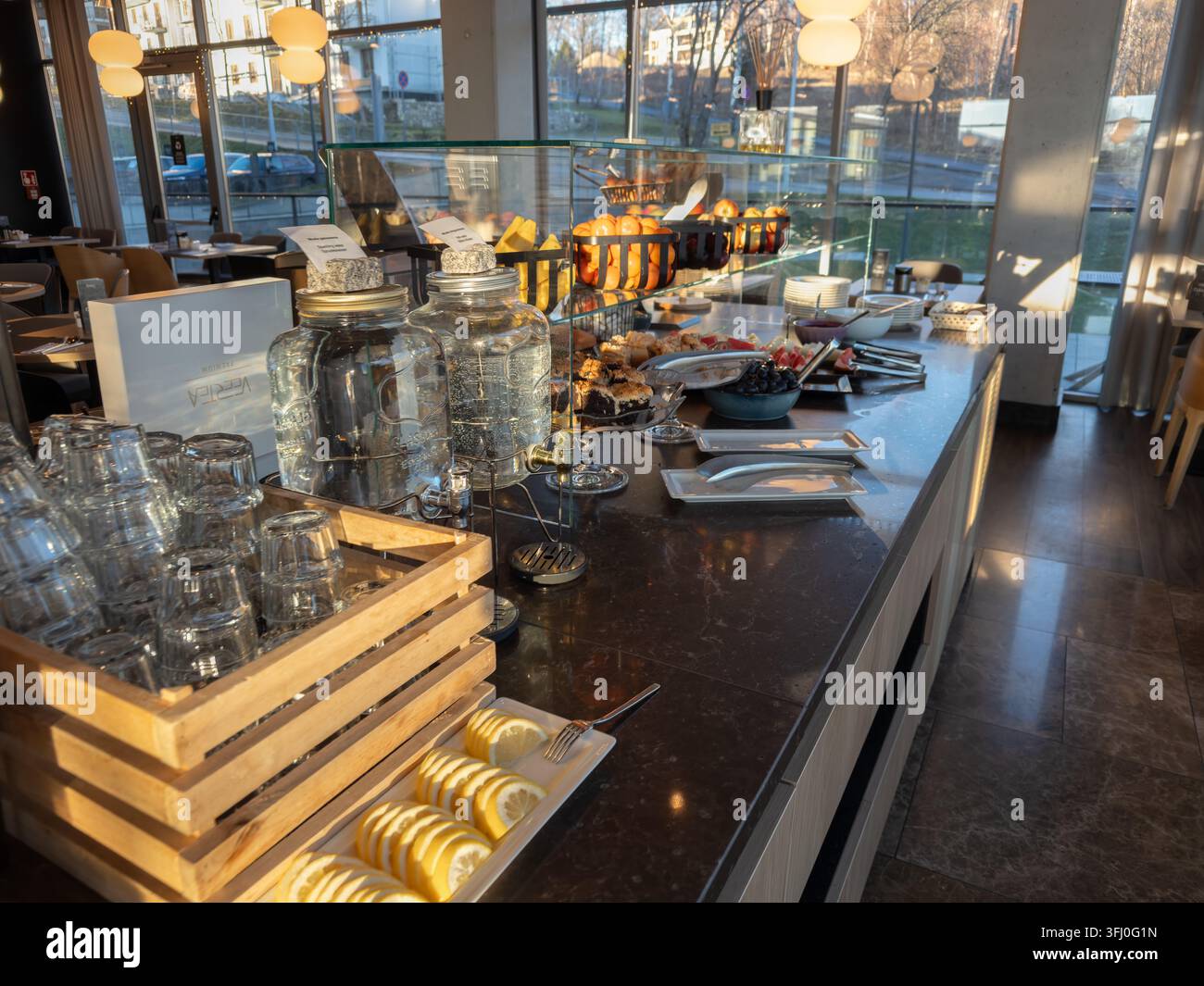 Ein organisierter Self-Service-Frühstücksbereich mit Glasgetränken und gesunden Snacks, die auf einem schlanken Tisch in einem sonnendurchfluteten Speisesaal arrangiert sind, der eine Prof... Stockfoto