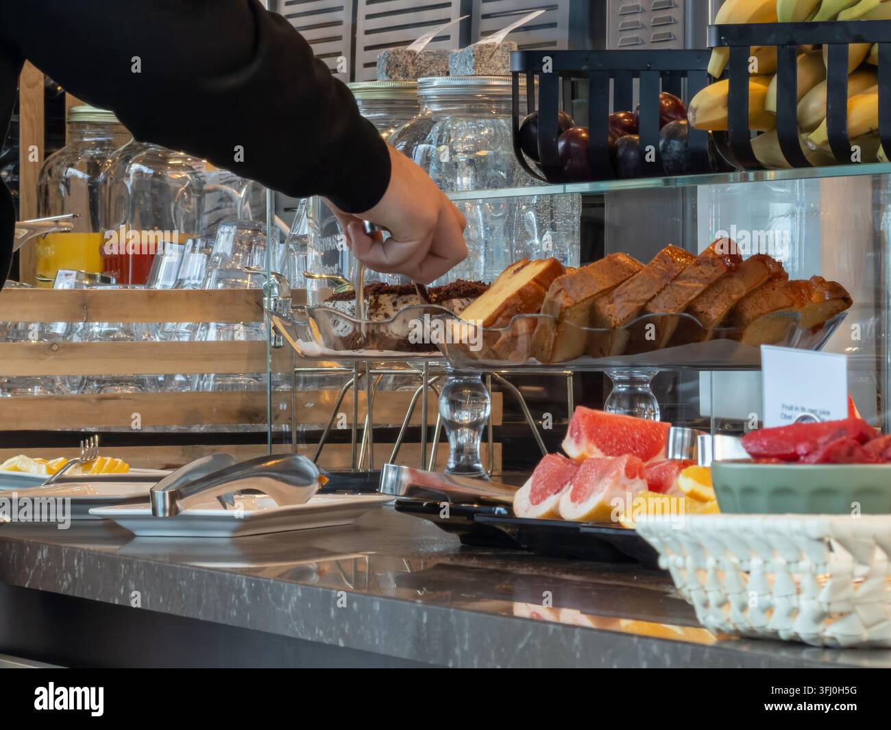 Eine Hand wählt mit einer Zange ein Stück Biskuitkuchen an einem farbenfrohen Frühstücksbuffet aus, das einen Gourmet-Hospitalitätsdienst für das Morgenessen präsentiert Stockfoto