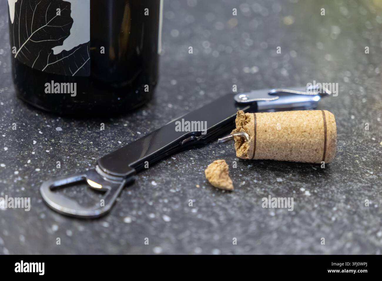Ein metallener Weinöffner liegt neben einem zerbrochenen Flaschenverschluss auf einer Granitarbeitsplatte, was einen chaotischen Öffnungsvorfall oder den Beginn eines lockeren Abends veranschaulicht Stockfoto