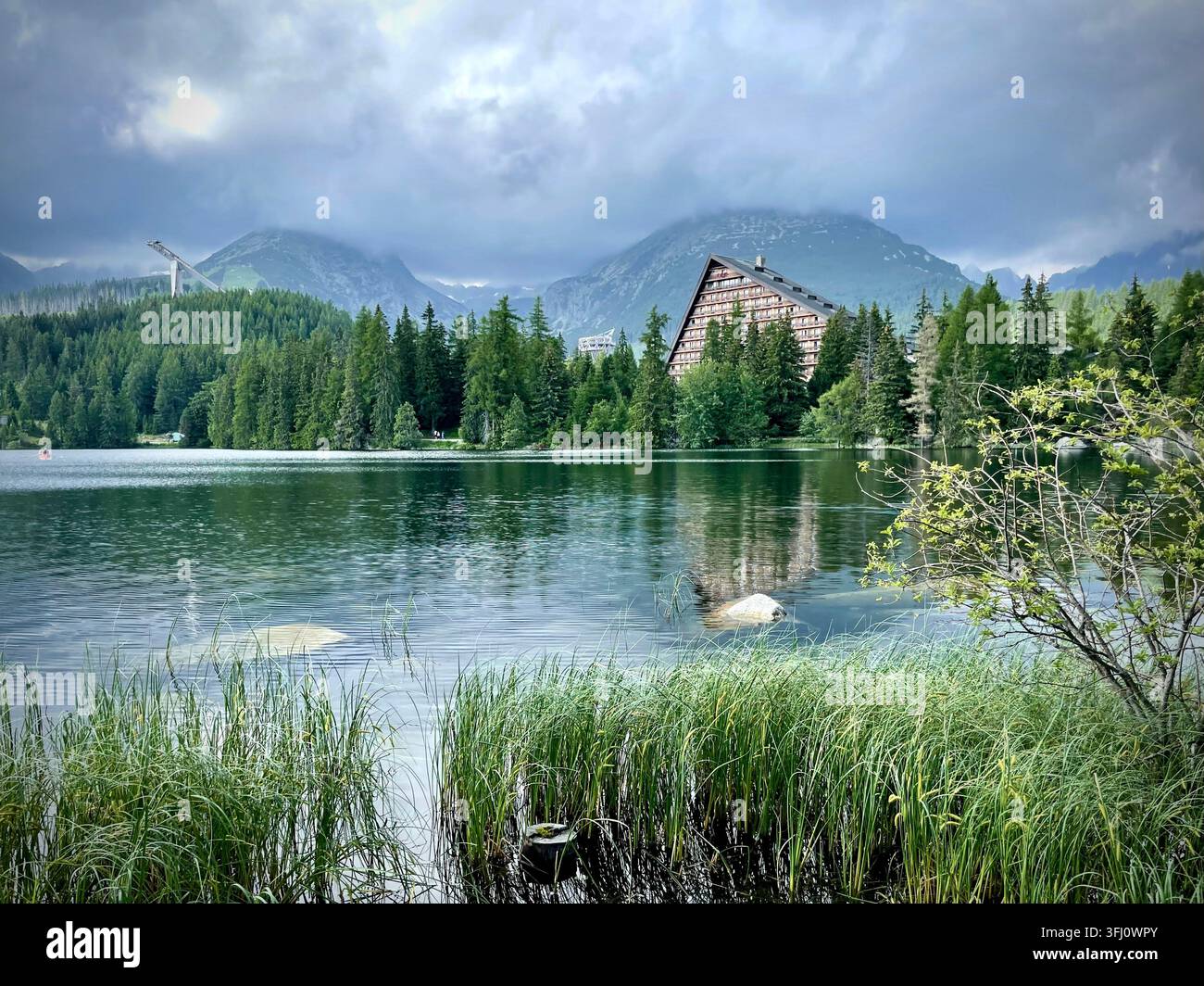 Panoramatische Ansicht von Touristen, die rote Boote auf dem Štrbské Pleso-See in der Hochtatra in der Slowakei rudern, mit Hotel Patria und nebligen Bergen im Hintergrund. Stockfoto