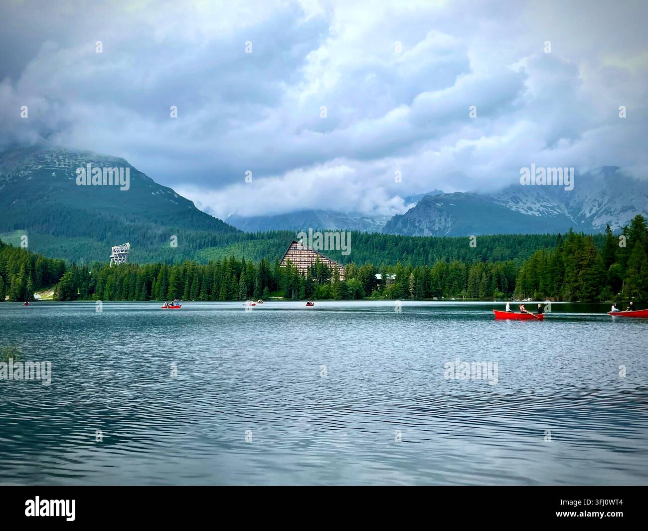 Panoramatische Ansicht von Touristen, die rote Boote auf dem Štrbské Pleso-See in der Hochtatra in der Slowakei rudern, mit Hotel Patria und nebligen Bergen im Hintergrund. Stockfoto