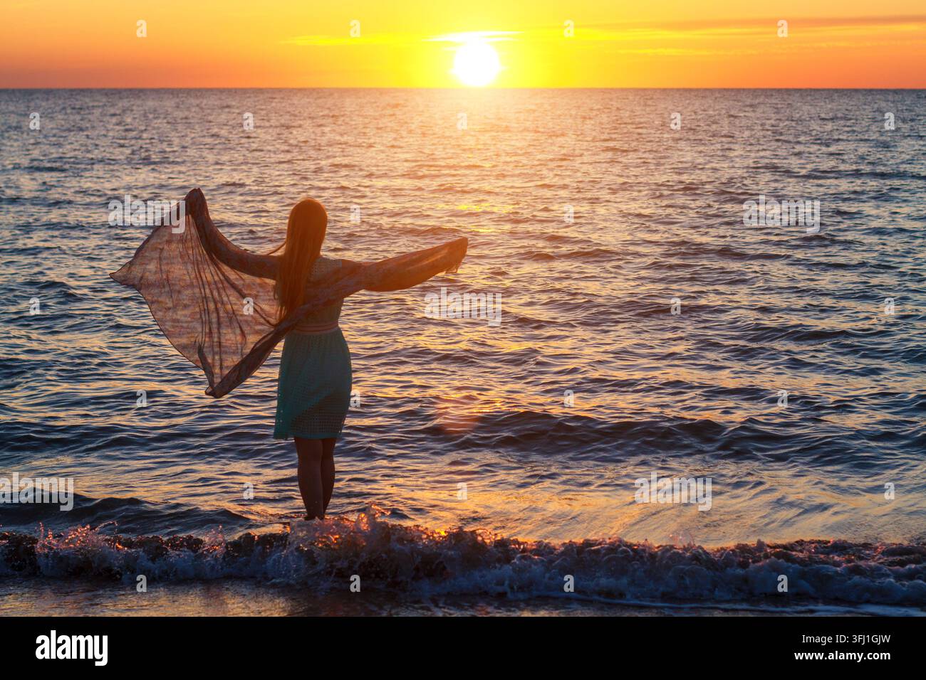 Frau in Mintgrünem Kleid mit breitem, fließendem Schal in Sea Sunset Bay Stockfoto