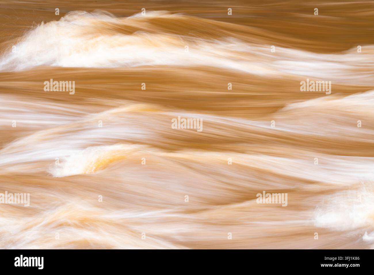 Seidige Wellen von schlammigem braunem Hochwasser fließen in Langzeitbelichtung über das Bild und bilden geschichtete Bänder der Bewegung mit sanften weißen Reflexen Stockfoto