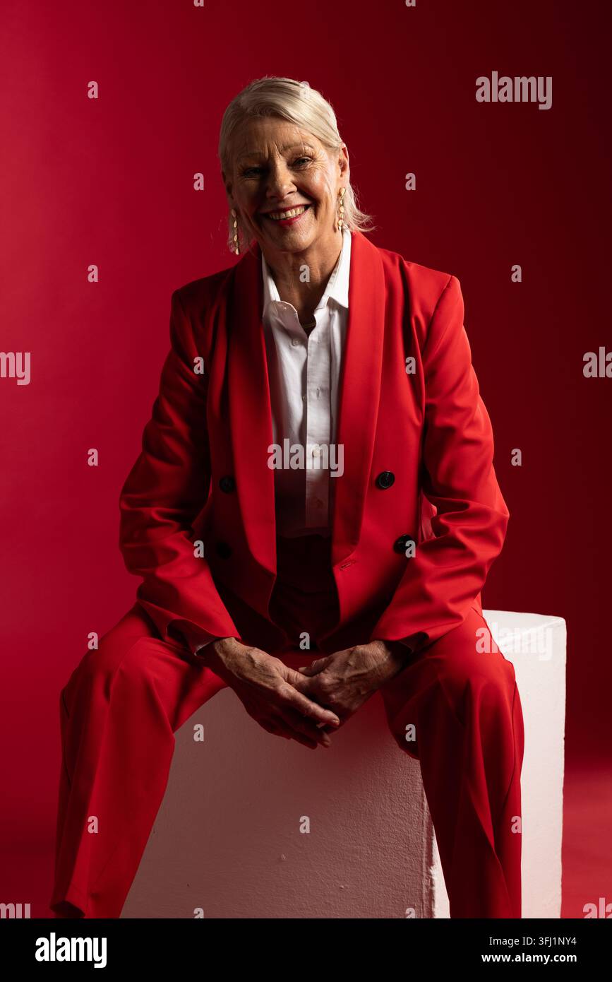 Seniorfrau lächelt und sitzt auf weißem Podest im Studio, trägt roten Blazer und Hose Stockfoto