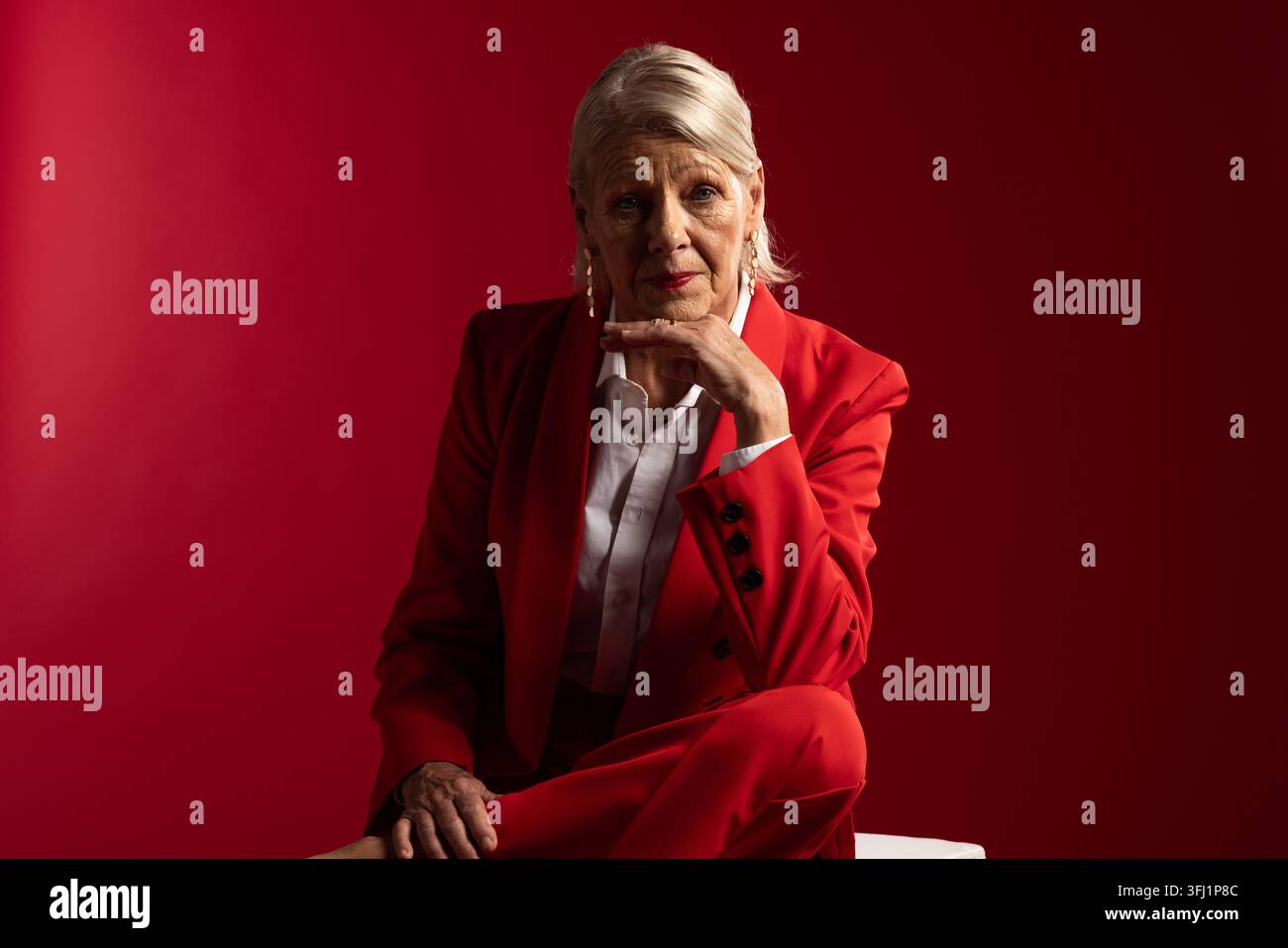 Seniorin sitzt auf weißem Würfel im Studio, trägt einen roten maßgeschneiderten Anzug, Goldohrringe, roter Hintergrund Stockfoto
