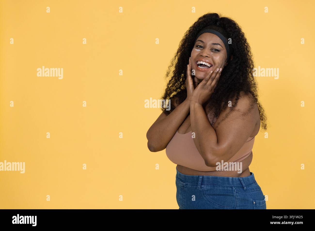 Frau, die Wangen berührt und lacht, beginnt lebhafte Werbung auf gelbem Hintergrund, der ein Outfit zeigt. Porträt, Studio, Jeans, cr Stockfoto