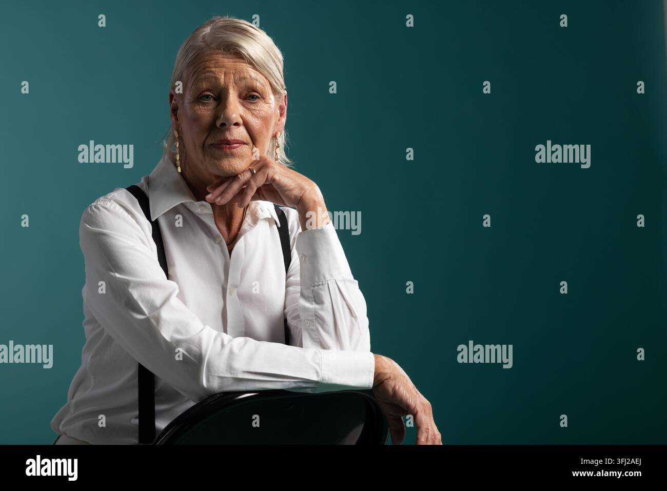 Seniorfrau sitzt auf einem Stuhl vor einer blaugrünen Studiokulisse und trägt ein weißes Hemd und Hosenträger Stockfoto