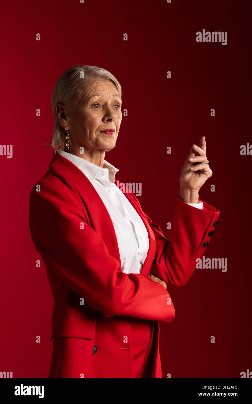 Seniorin posiert im Studio, trägt roten Blazer und Hose, weißes Hemd, Tropfenohrringe und Ring Stockfoto