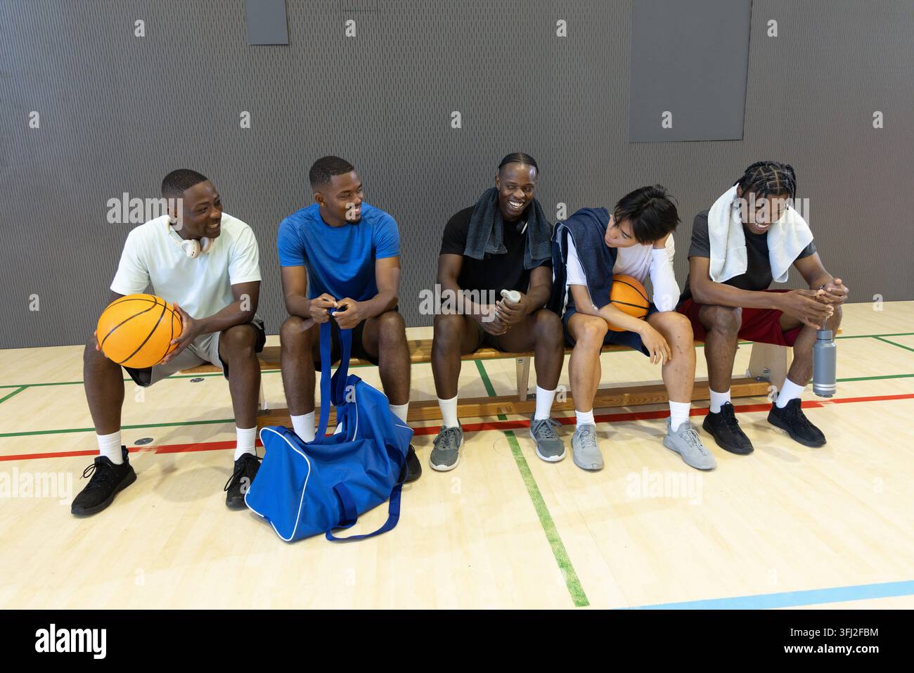 Verschiedene männliche Mannschaftskameraden sitzen im Fitnessstudio in Trikots, halten Basketbälle und Duffel. Athleten, Kameradschaft, Court, Hartholz, Training, Zahnrad Stockfoto