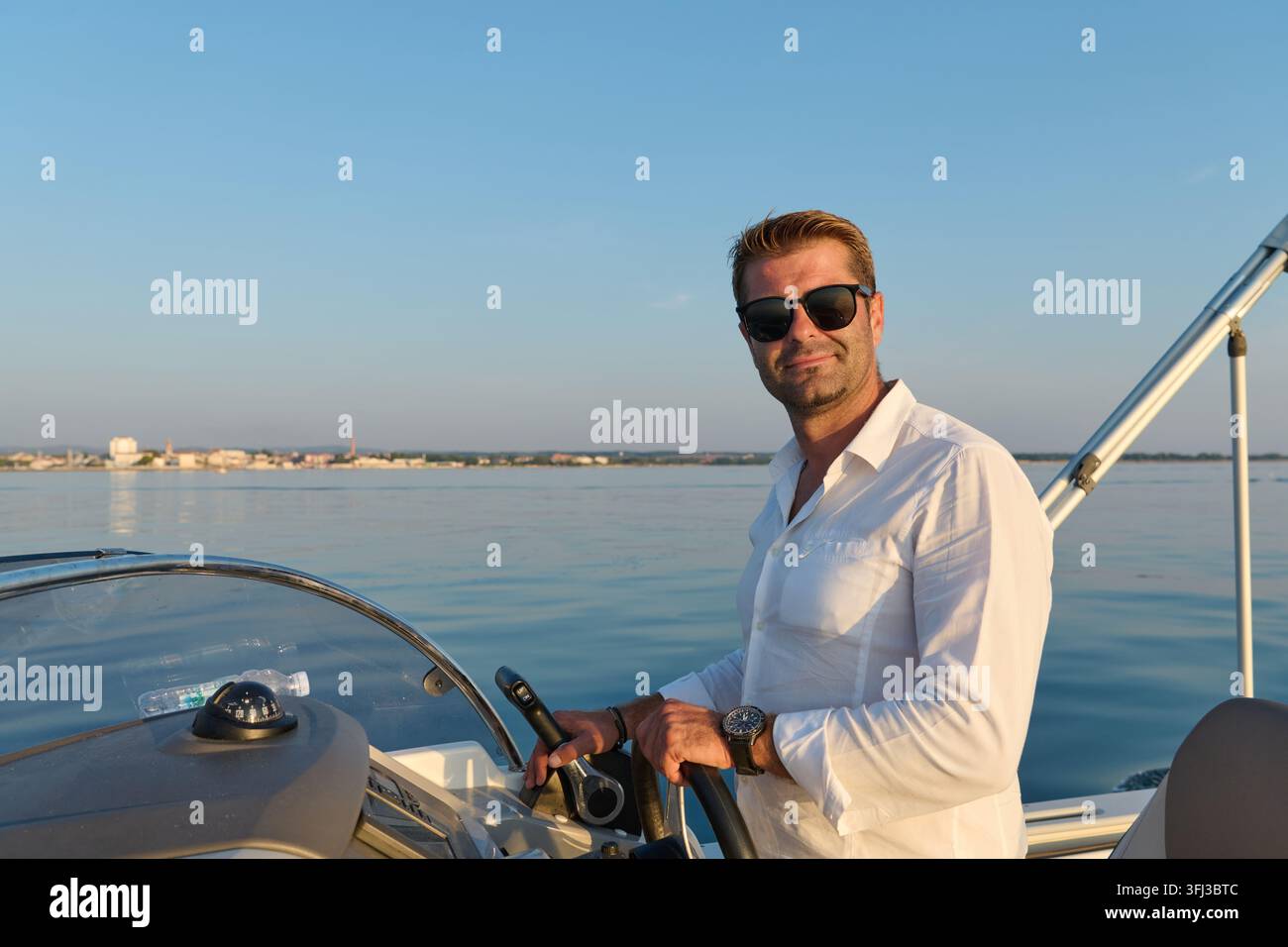 Ein entspannter Mann in einem weißen Leinenhemd und Sonnenbrille steuert ein Motorboot vor der Küste von Umag zur goldenen Stunde, ruhige Adriawasser und ferne Küste e Stockfoto