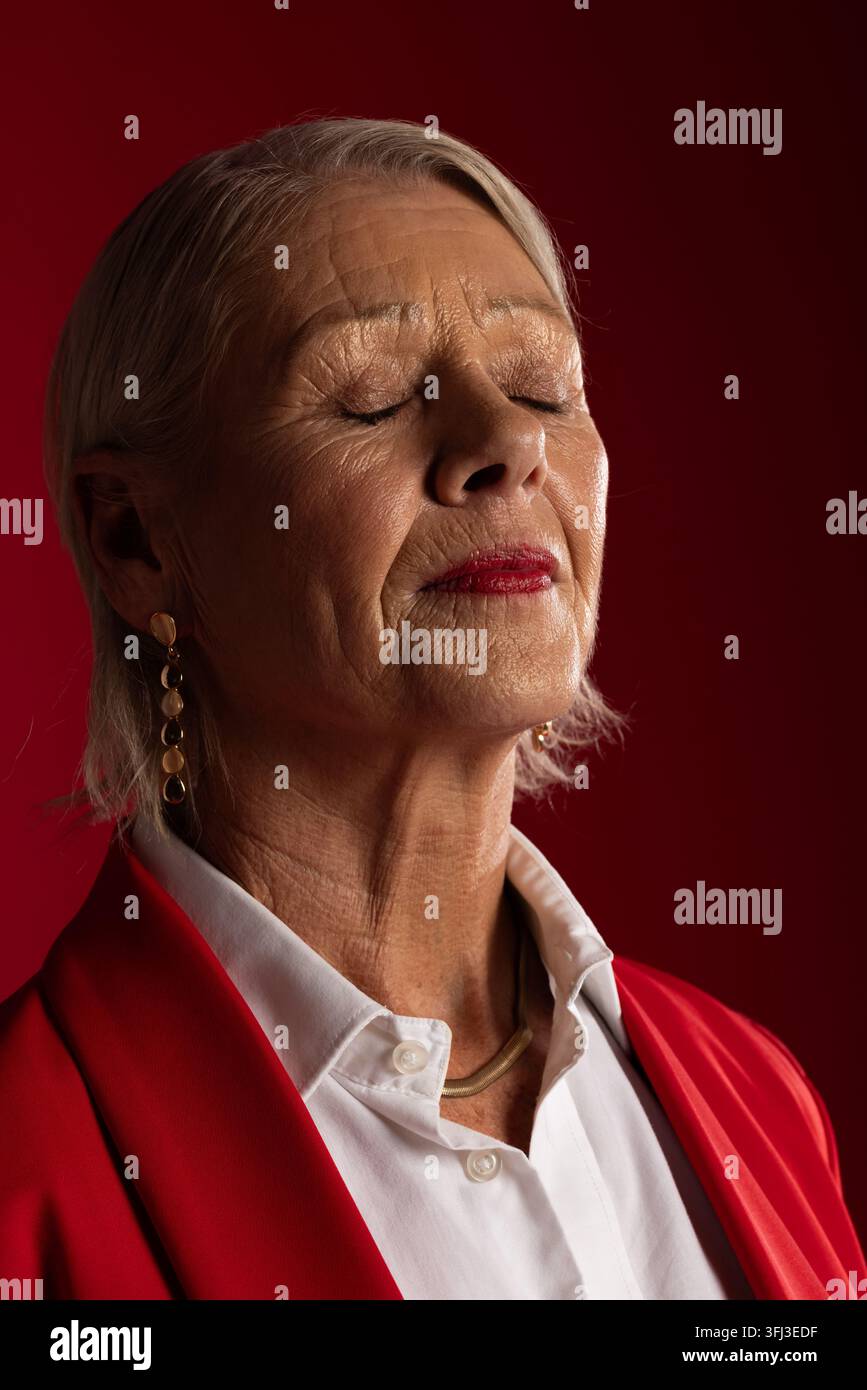 Seniorin schließt die Augen, neigt den Kopf nach oben im Studio, trägt roten Blazer, Goldschmuck, roten Lippenstift Stockfoto