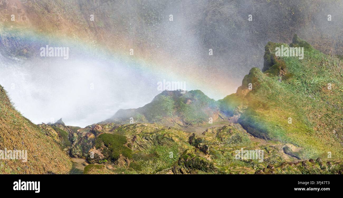 Regenbogen, der sich über das moosbedeckte felsige Gelände unter dem Wasserfall spannt Stockfoto