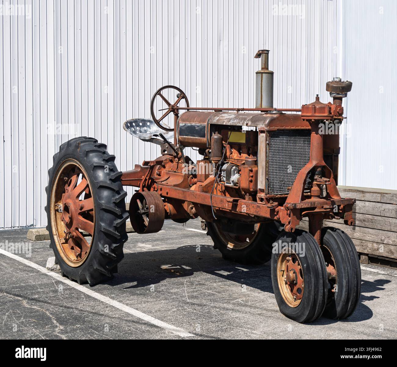 Rostiger Vintage-Traktor, der auf Beton geparkt ist. Große schwarze Reifen und Metallteile zeigen Abnutzungsspuren. Weißer vertikaler Wandverkleidung bildet den Hintergrund. Sonnenlicht wirft Schatten Stockfoto