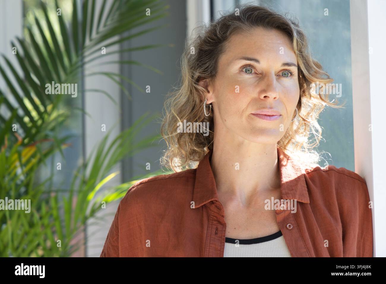 Mitte-alte Frau steht nahe am Fenster, Sonnenlicht umrahmt lockiges Haar, rostfarbenes Hemd und Pflanze. Weiblich, Porträt, Interieur, gelassen Stockfoto