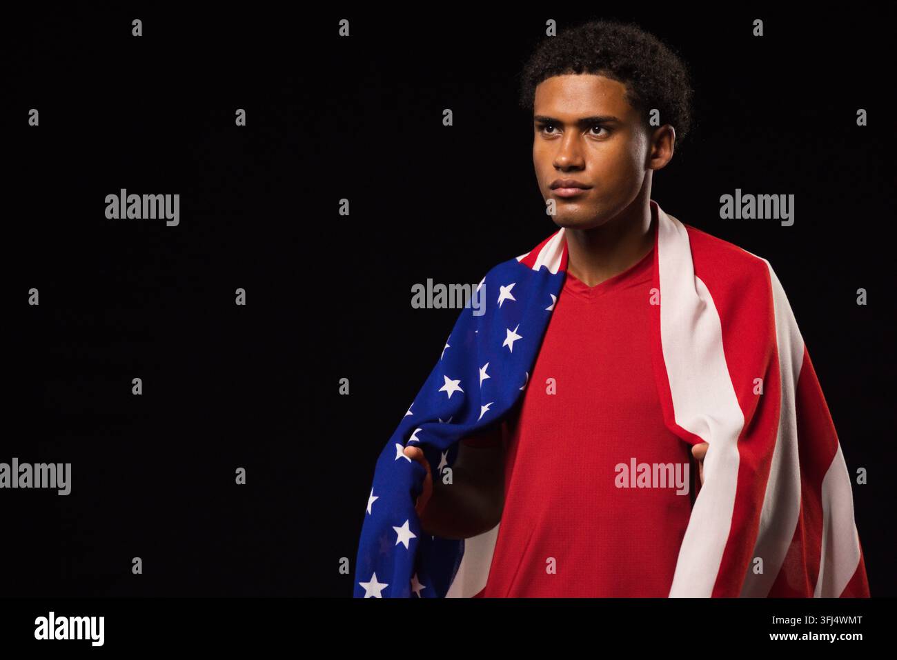 Afroamerikaner steht im Studio, trägt rotes T-Shirt und drapiert die US-Flagge, Kopierraum. Porträt, stoisch, patriotisch, Scheinwerferlicht Stockfoto