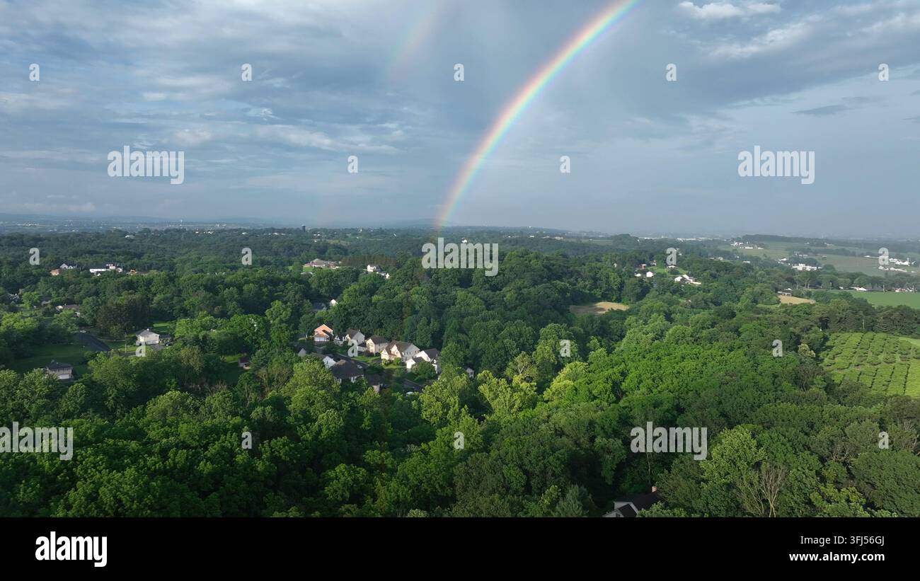 Amerikanische Siedlung und Wohngebiet zwischen grünen Bäumen auf dem Hügel. Ländlicher Bereich einer amerikanischen Stadt. Schöner Regenbogenbogen am Himmel. Luftbild mit großer Brennweite. Stockfoto