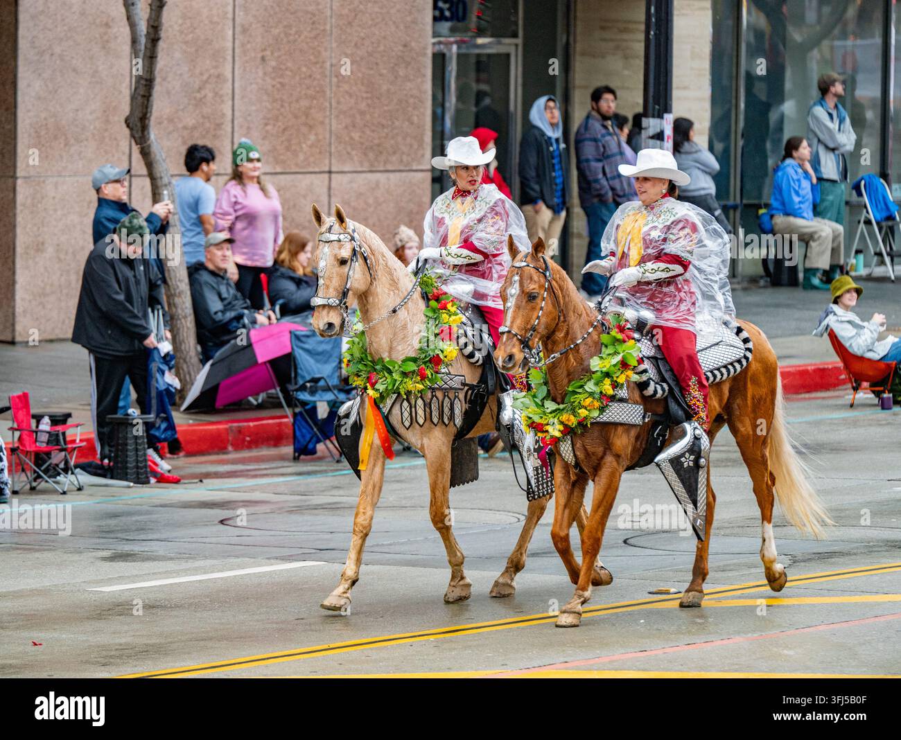 Reiter in Regenponchos reiten Pferde in Blumengirlanden die nassen Straßen während der 137. Rose Parade hinunter. Stockfoto