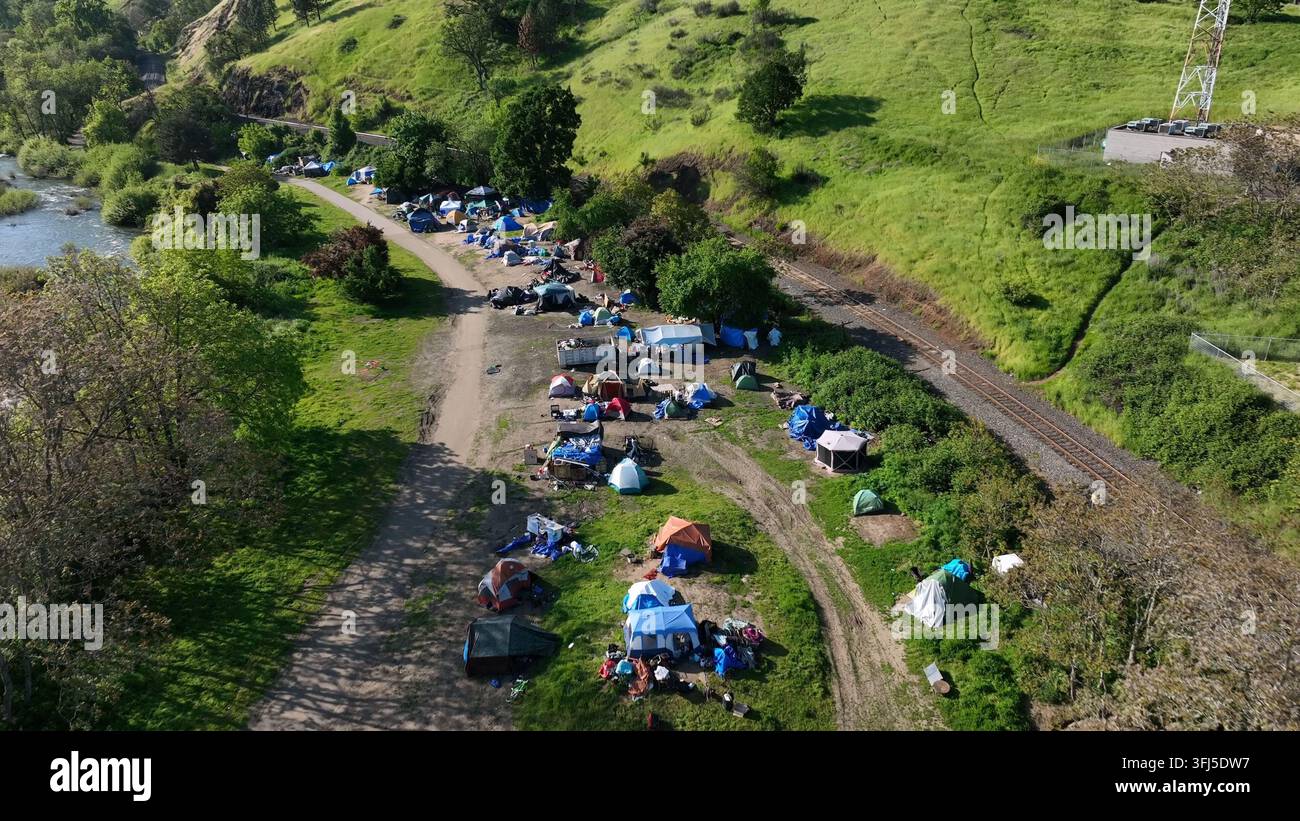 Obdachlosenlager mit bunten Zelten und blauen Planen breitet sich entlang von Schotterwegen neben dem Fluss unter einem üppigen grünen Hügel an einem sonnigen Frühlingstag in Roseb aus Stockfoto