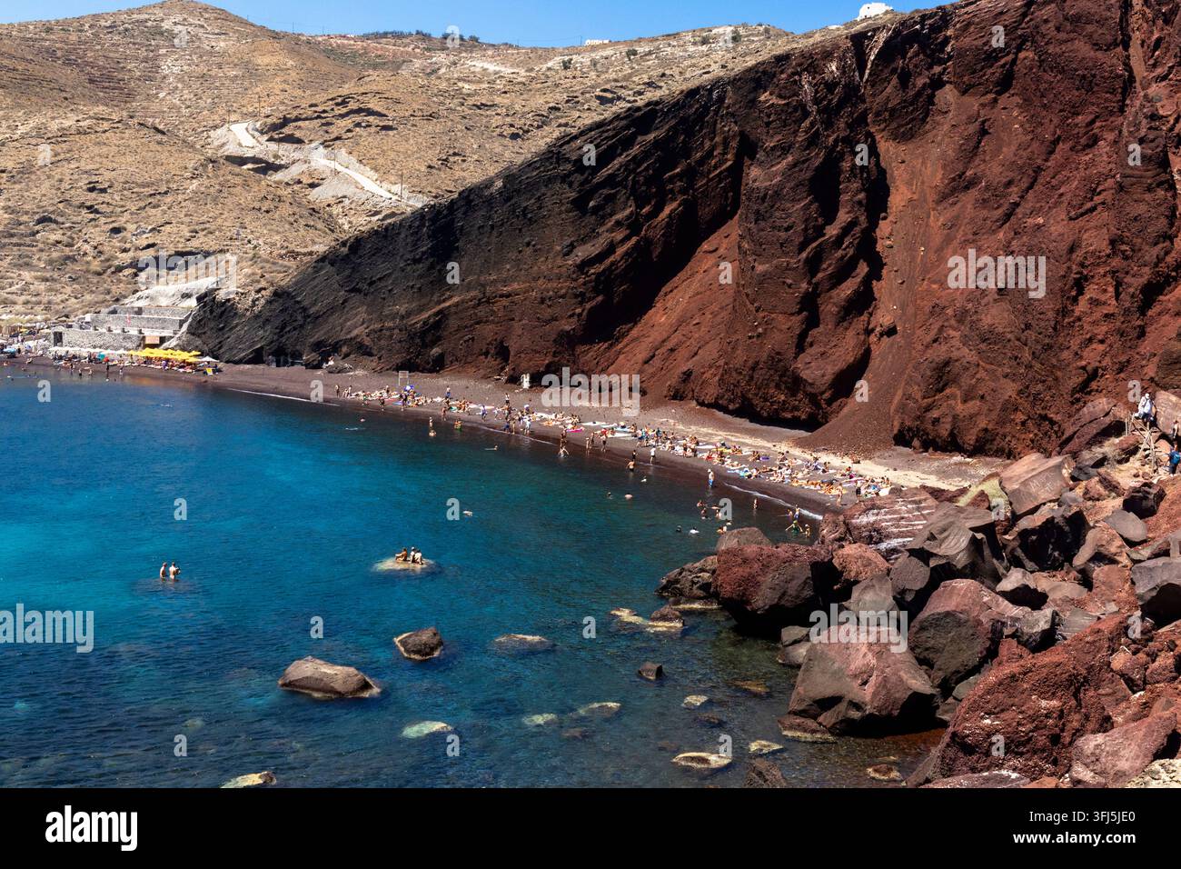 Panoramablick auf Red Beach (Kokkini Paralia) in Santorini, Griechenland, mit dramatischen roten Vulkanfelsen, dunklem Sand, türkisfarbenem Wasser und Touristen e Stockfoto