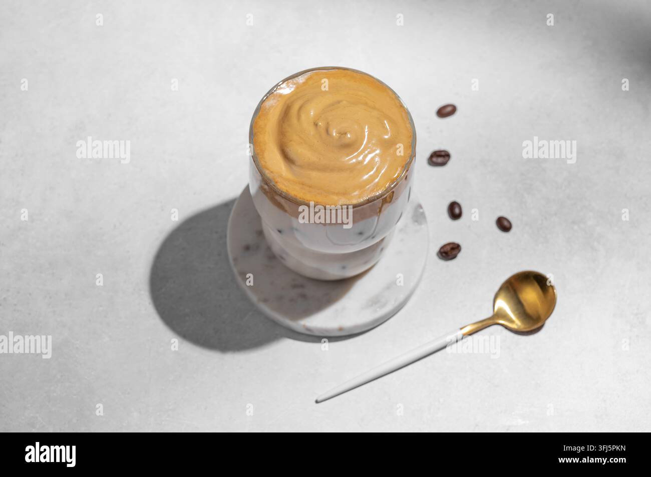 Dalgona-Kaffee. Geschlagener Instant-Eiskaffee in einem Glas mit Kaffeebohnen, Löffel und Schatten auf einem grauen Betonhintergrund. Trendiges süßes Dessertkonzept Stockfoto