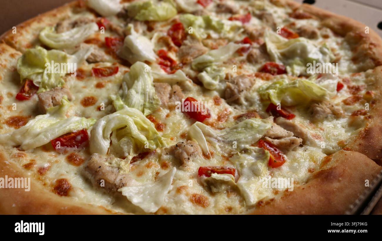Eine appetitliche Caesar-Pizza auf einem luftigen Teig mit einem goldenen Rand. Sie ist belegt mit zart geschmolzenem Käse, saftigen Hühnerfiletstücken, reifen Kirschtomaten Stockfoto