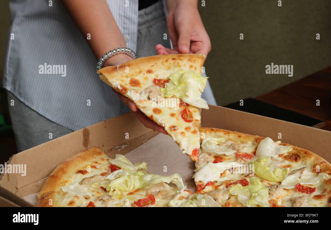 Eine Person nimmt eine appetitliche dreieckige Pizzascheibe aus einem Karton; die Pizza hat einen dicken Rand, weiße Soße, geschmolzenen Käse, Hühnchen Stockfoto