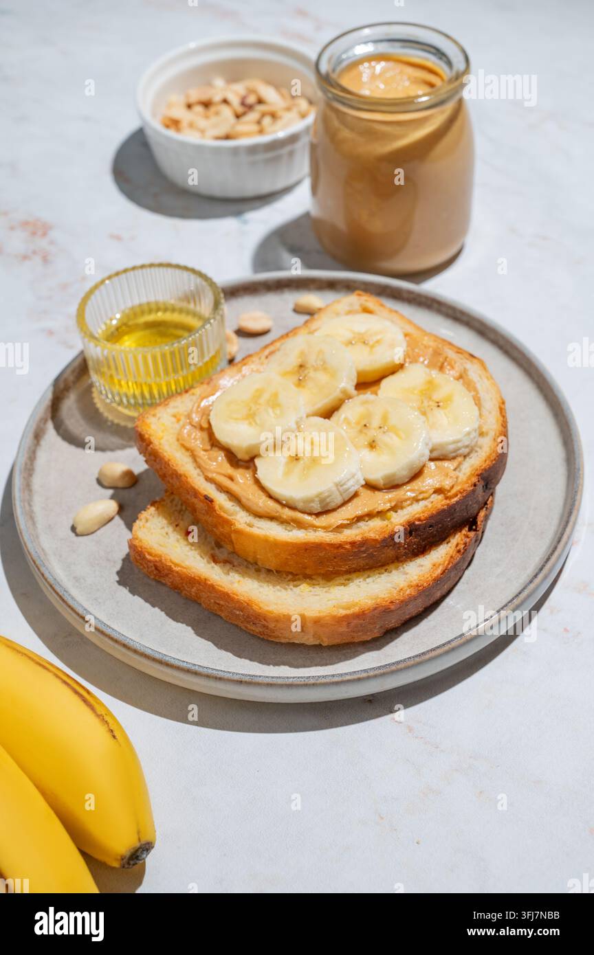 Erdnussbutter-Toast und Banane auf einem Teller auf einem Marmorhintergrund mit Früchten, Nüssen, Honig und Morgenschatten. Konzept von süßer Ernährung und gesunden Snacks Stockfoto