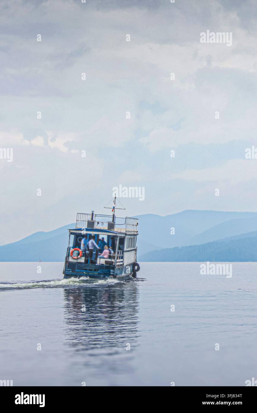 Passagierboot mit Menschen an Bord kreuzt über ruhiges Wasser mit Bergen im Hintergrund und hinterlässt eine sichtbare Spur. Malerische Wasserfahrt, zu Stockfoto