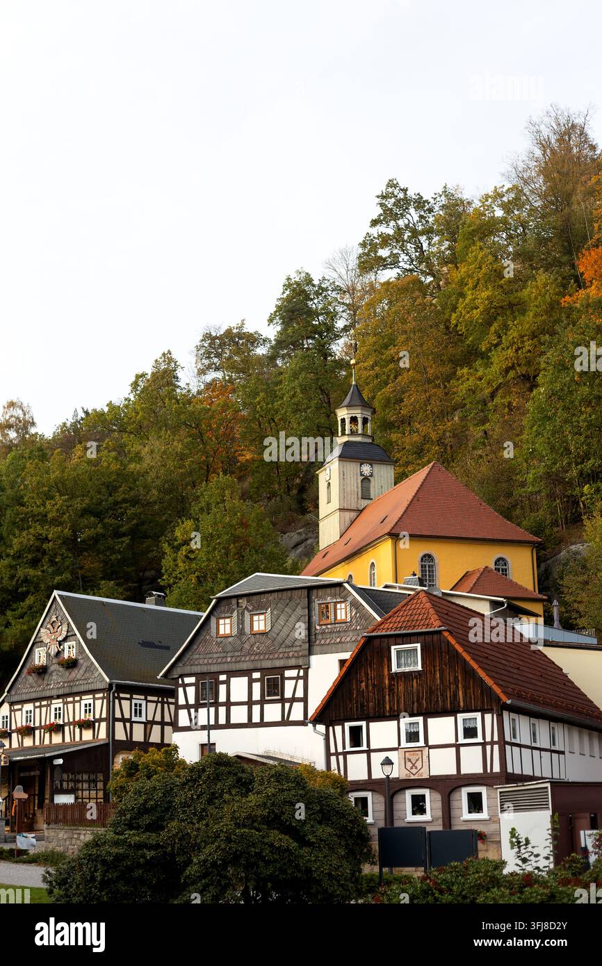 Pitoreskes Dorf mit historischen Fachwerkhäusern und einem gelben Kirchengebäude, das am Fuße eines felsigen Hügels mit Herbstbäumen in Oybin eingebettet ist Stockfoto