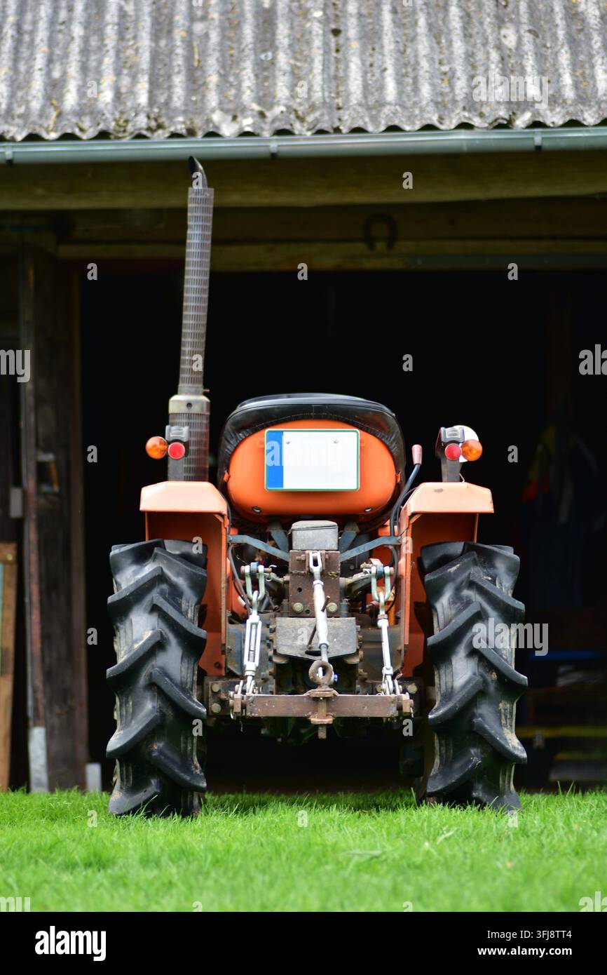Rückansicht eines kompakten orangefarbenen Traktors, der vor einer rustikalen Holzbarn geparkt ist. Traditionelle landwirtschaftliche Maschinen und authentische Bauernhoflandschaft Stockfoto