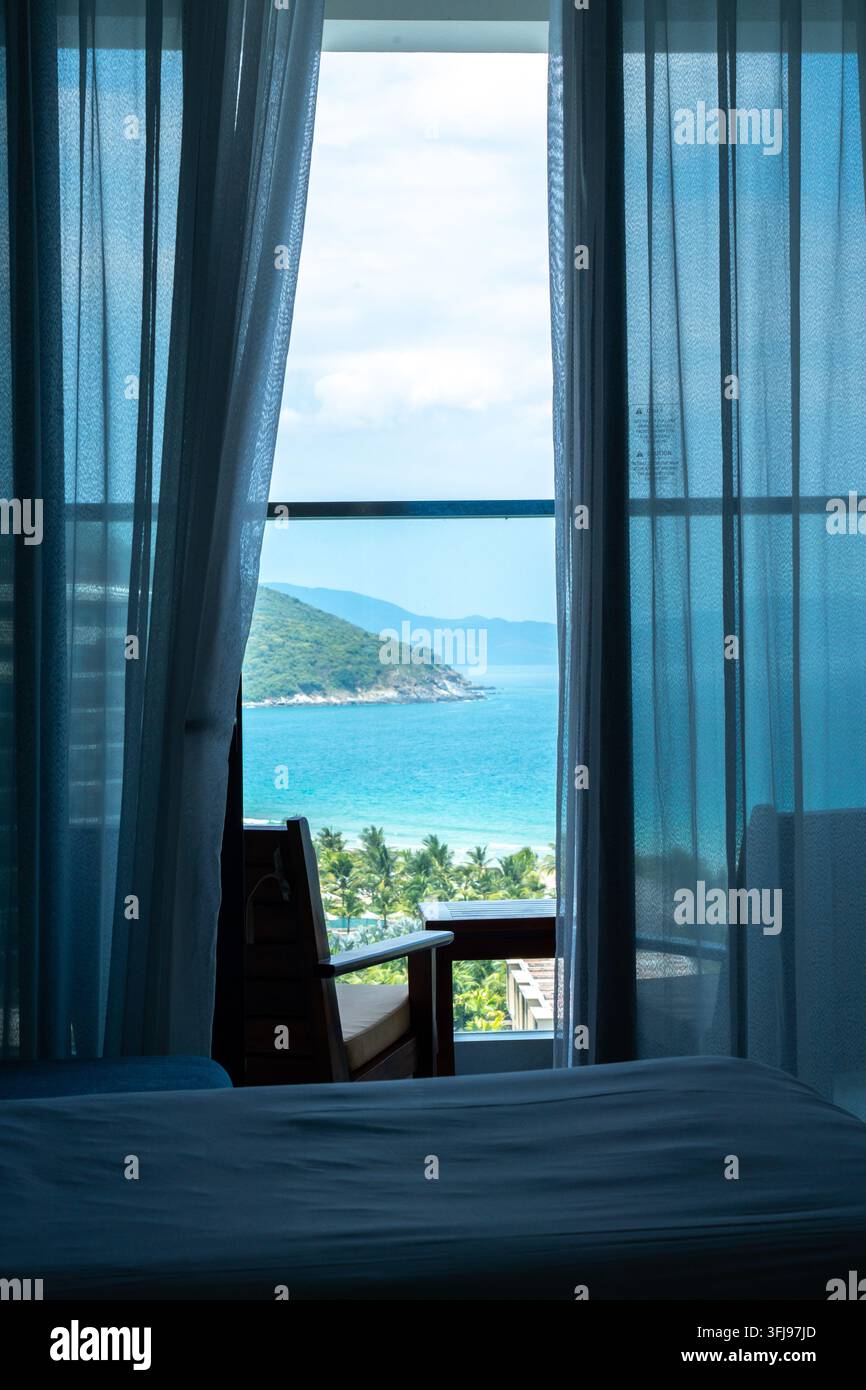 Serene Küstenaussicht durch Vorhänge: Ein ruhiger Rückzugsort in die Natur mit atemberaubendem Meer- und Bergpanorama, sichtbar von einem gemütlichen Innenraum. Stockfoto