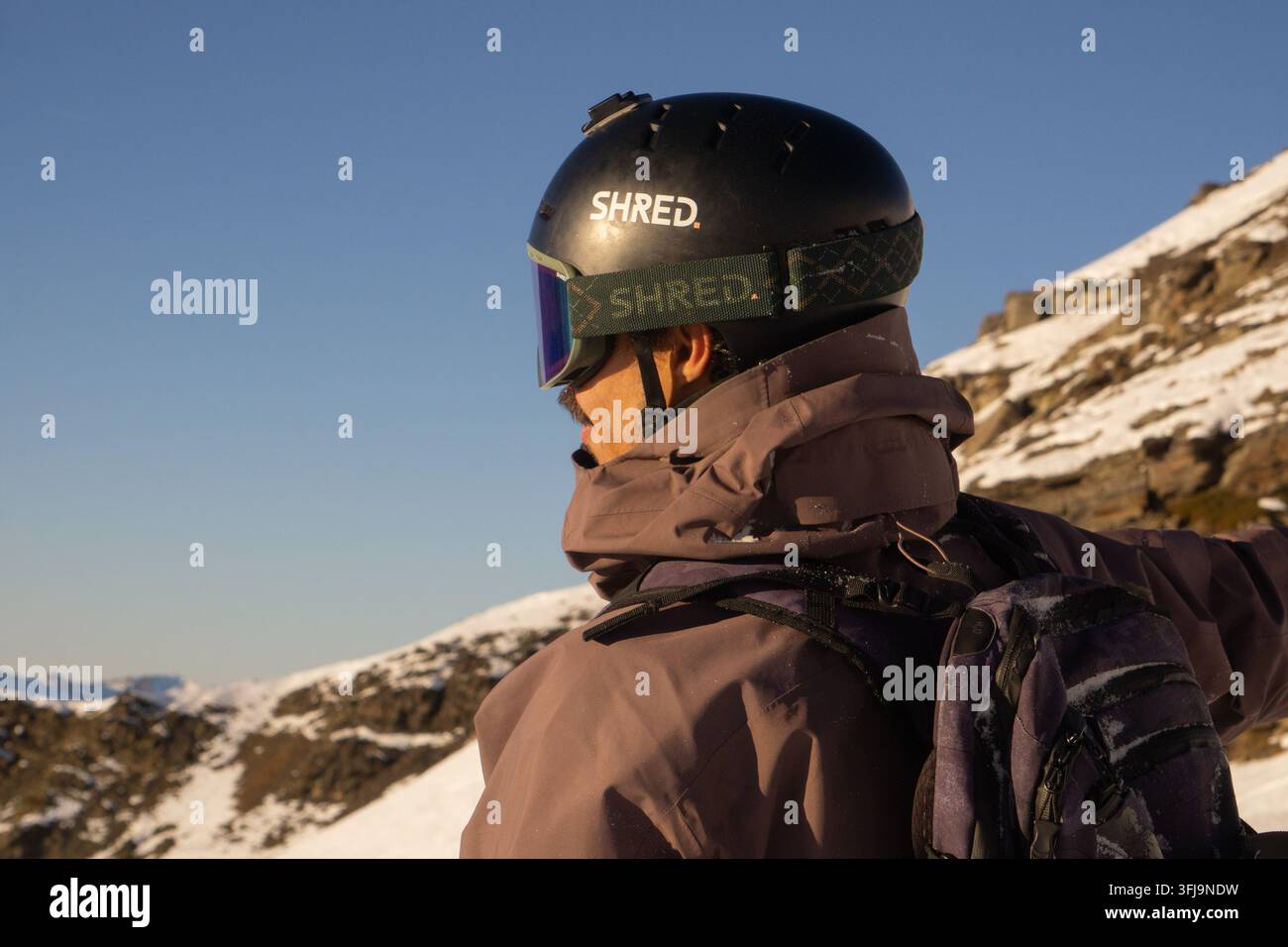 Person, die einen Helm, gespiegelte Schutzbrille und einen Rucksack trägt, neben schneebedeckten, felsigen Hängen in Val Thorens, Drei Täler, französische Alpen, Frankreich. Stockfoto