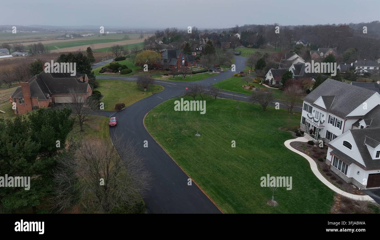 Luftaufnahme einer ruhigen, vornehmen amerikanischen Vorstadtgegend mit großen Häusern, geschwungenen Straßen, offenen Rasenflächen und umgebendem Ackerland an einem bewölkten Spätnachmittag Stockfoto