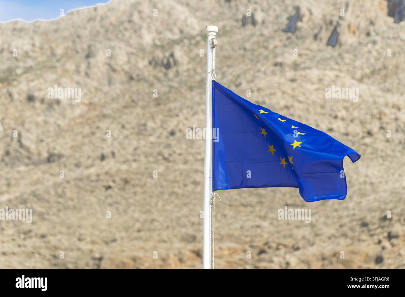Eine blaue Sternenflagge weht gegen den felsigen Halki-Hügel, ihre helle Farbe setzt sich in klarem Inselhimmel und warmem Nachmittagslicht ab. Stockfoto
