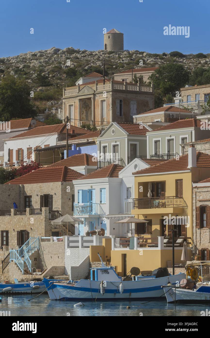Farbenfrohe Halki-Häuser erheben sich über ruhigem Wasser und felsigem Land, während ein hoher Kirchturm über kleinen Booten im klaren Insellicht wacht. Stockfoto