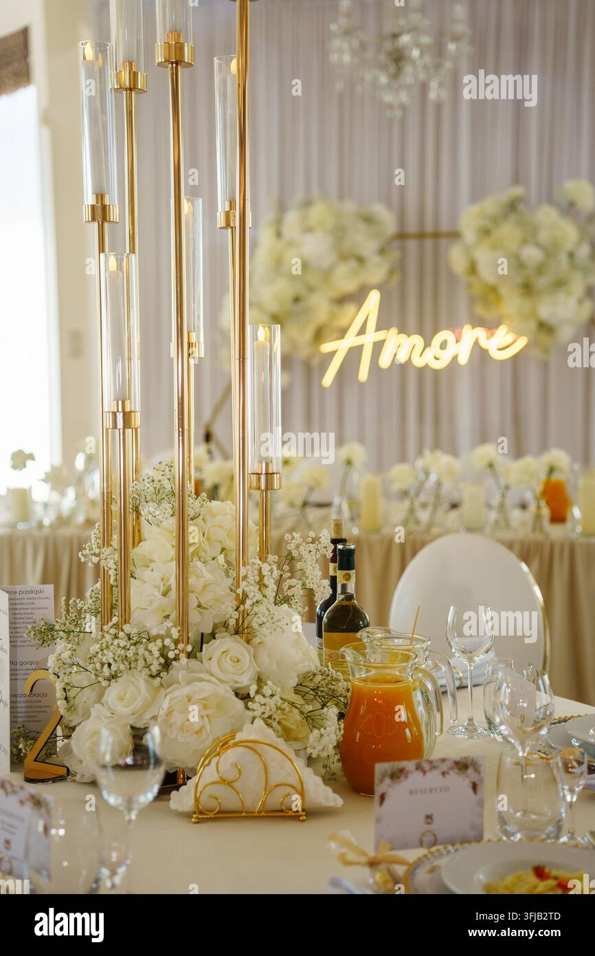 Eleganter Hochzeitstisch-Mittelstück mit hohen goldenen Kerzenständern und weißen Blumen bei einer romantischen Empfangsparty mit einem leuchtenden dekorativen Neonschild Stockfoto
