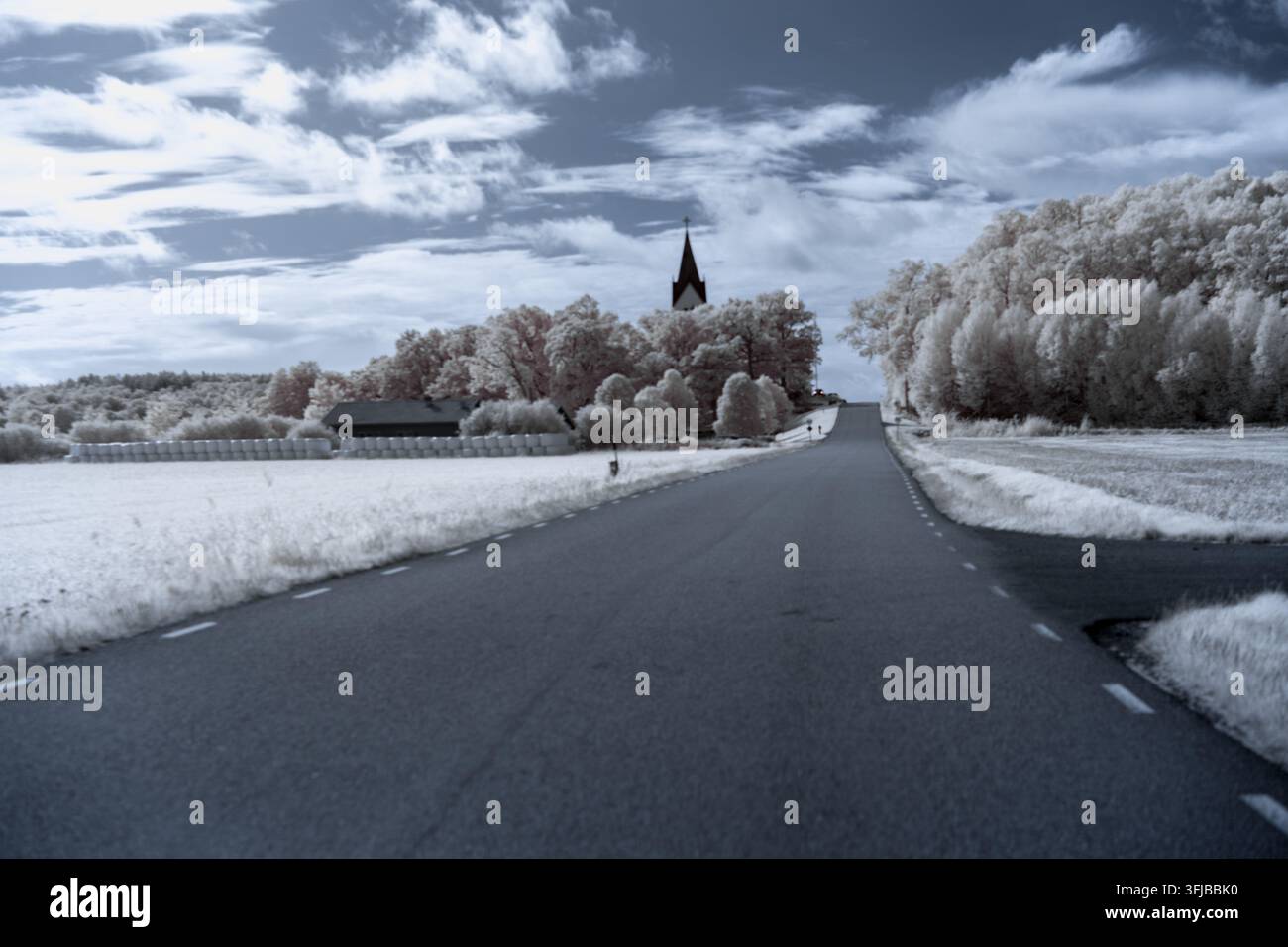 Eine Straße erstreckt sich in die Ferne mit einer Kirche am Ende. Bäume säumen die Seiten der Straße und Felder sind vorhanden. Diese Szene verwendet Infrarot Stockfoto