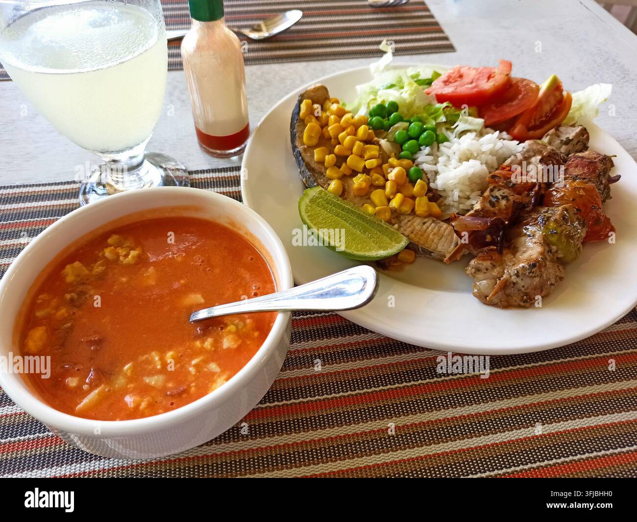 Nahaufnahme eines herzhaften Mahls mit gegrillten Fleischspießen, weißem Reis, gelbem Mais, grünen Erbsen, geschnittenen Tomaten, Limettenscheibe auf weißem Teller. Schüssel mit roter s Stockfoto