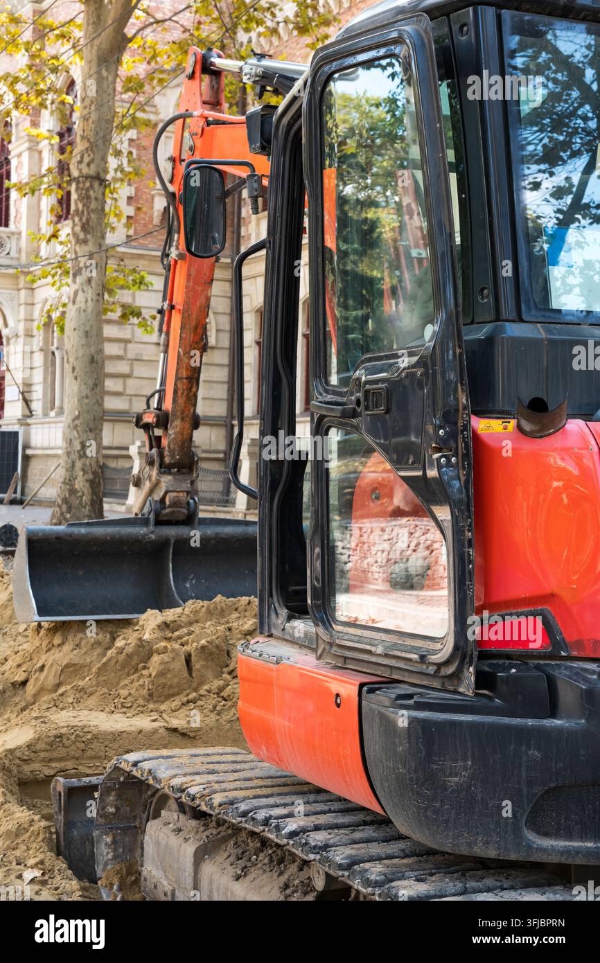Ein kleiner orangefarbener Raupenbagger, der bei Grabungsarbeiten während der Straßenreparatur und der Versorgungseinrichtung auf einer Stadtstraße arbeitet. Stockfoto