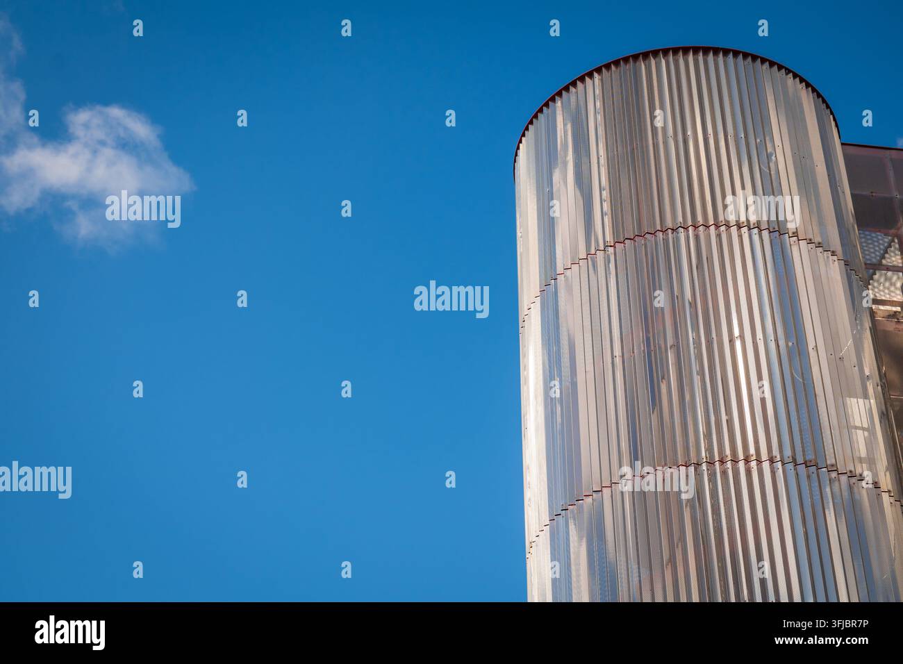 Eine moderne Metallzylinderfassade an einem zeitgenössischen Gebäude erhebt sich vor einem klaren blauen Himmel, das perforierte Netz erzeugt reflektierende Muster, starke Ver Stockfoto