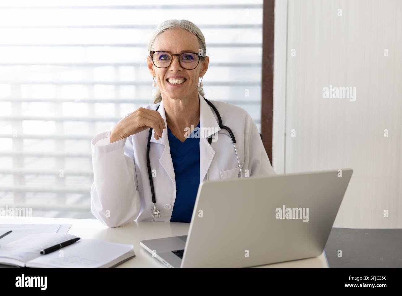 Porträt einer mittelalten Ärztin, die an einem Schreibtisch mit Laptop sitzt Stockfoto