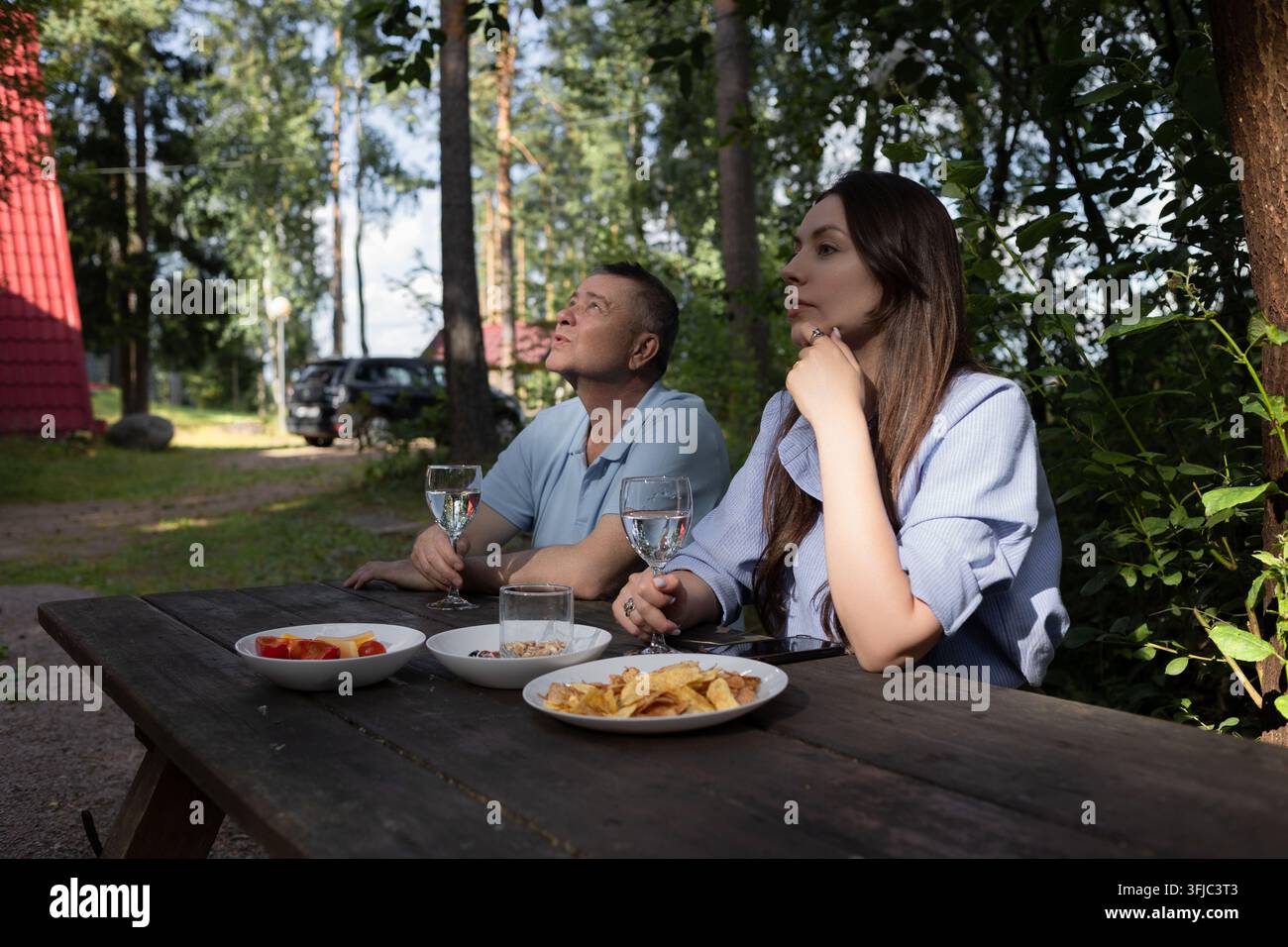 Vater und erwachsene Tochter an einem Picknicktisch auf dem Land, die gemeinsam etwas beobachten. Snacks, Chips und Getränke auf einem groben Holztisch unter pi Stockfoto