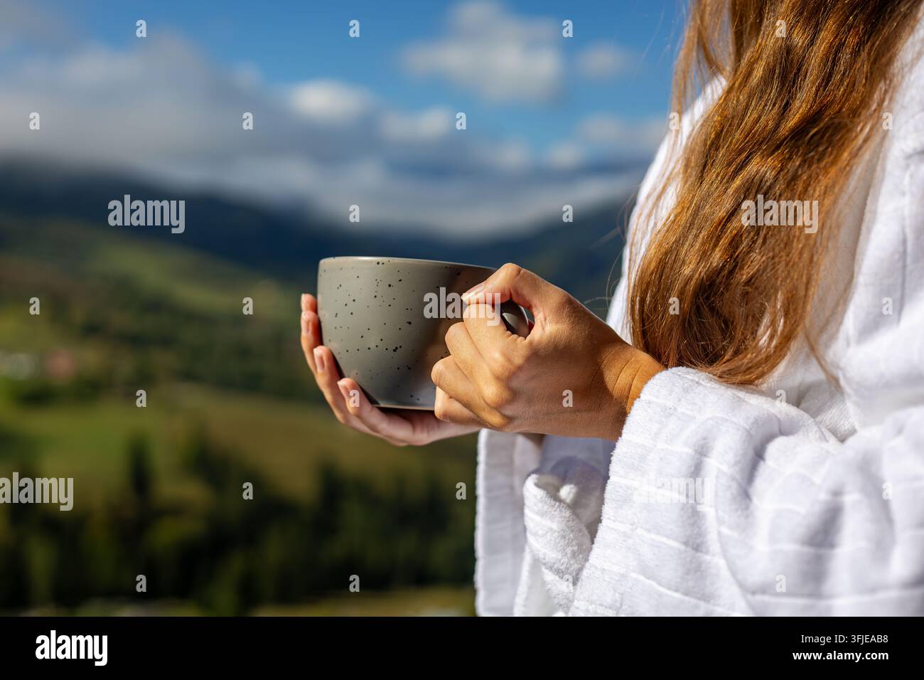 Frau hält eine Tasse vor einer gebirgigen Talfolge im Hintergrund. Eine Frau in einem weißen Bademantel hält eine graue Tasse gegen einen weich verschwommenen Bergtalhintergrund unter einem blauen Stockfoto